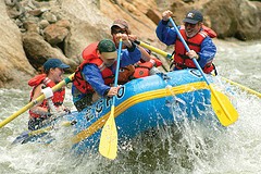Several people in a blue rafting boat paddling with whitewater pushing the boat up