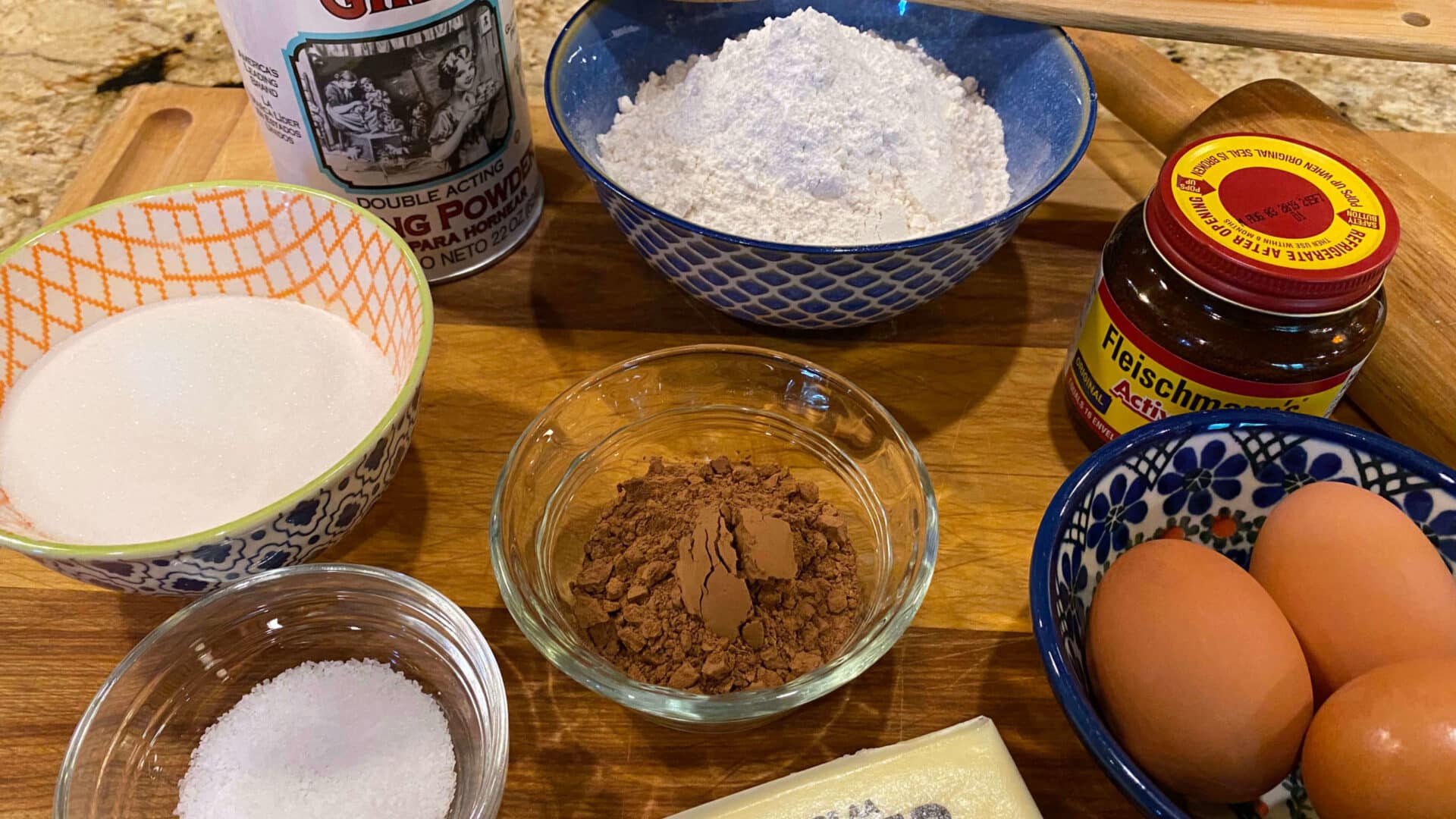 Baking Ingredients of flour, baking powder, yeast, cocoa, sugar, salt, eggs and butter, with a wooden rolling pin and wooden spoon