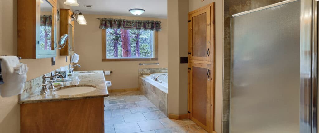 A large bathroom with double sinks in a granite countertop, a shower and jetted tub, and a large window with pine trees outside.