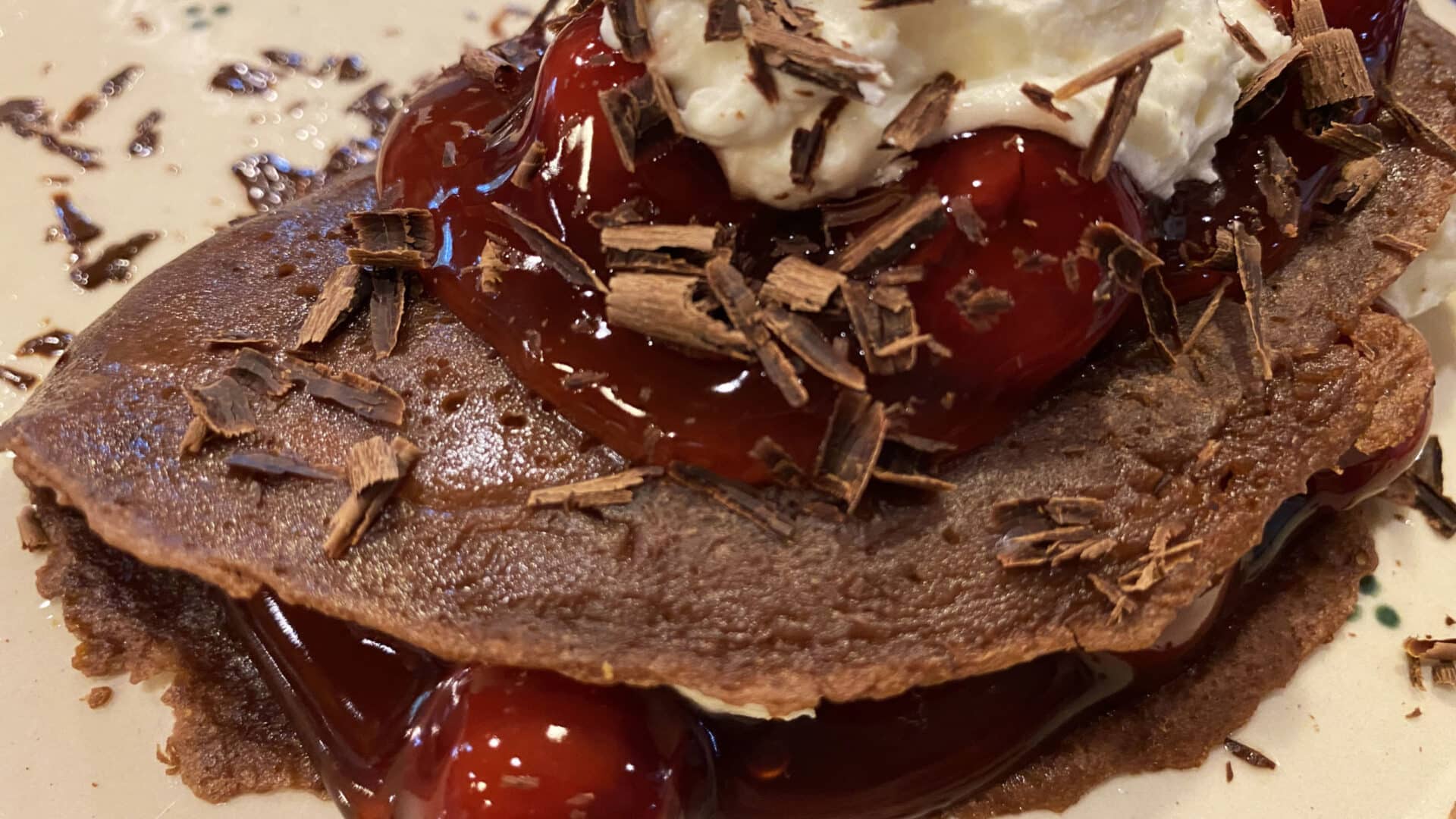 Chocolate crepes with cherry pie filling and cream in between and on top, garnished with shaved chocolate curls