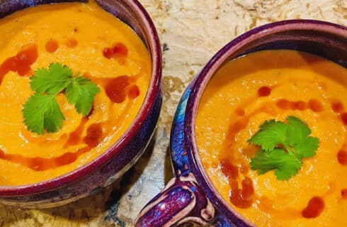 Orange Carrot-Coconut Soup drizzed with a red Thai sauce and garnished with green cilantro leaves in purple soup bowls