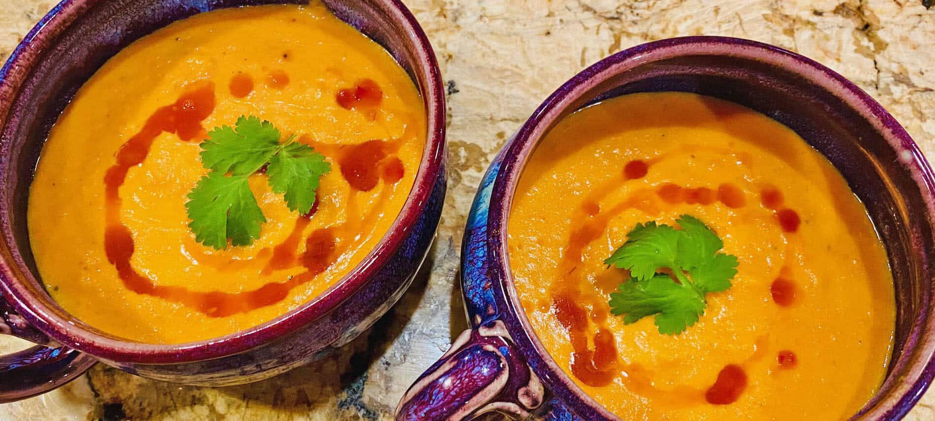 Orange Carrot-Coconut Soup drizzed with a red Thai sauce and garnished with green cilantro leaves in purple soup bowls