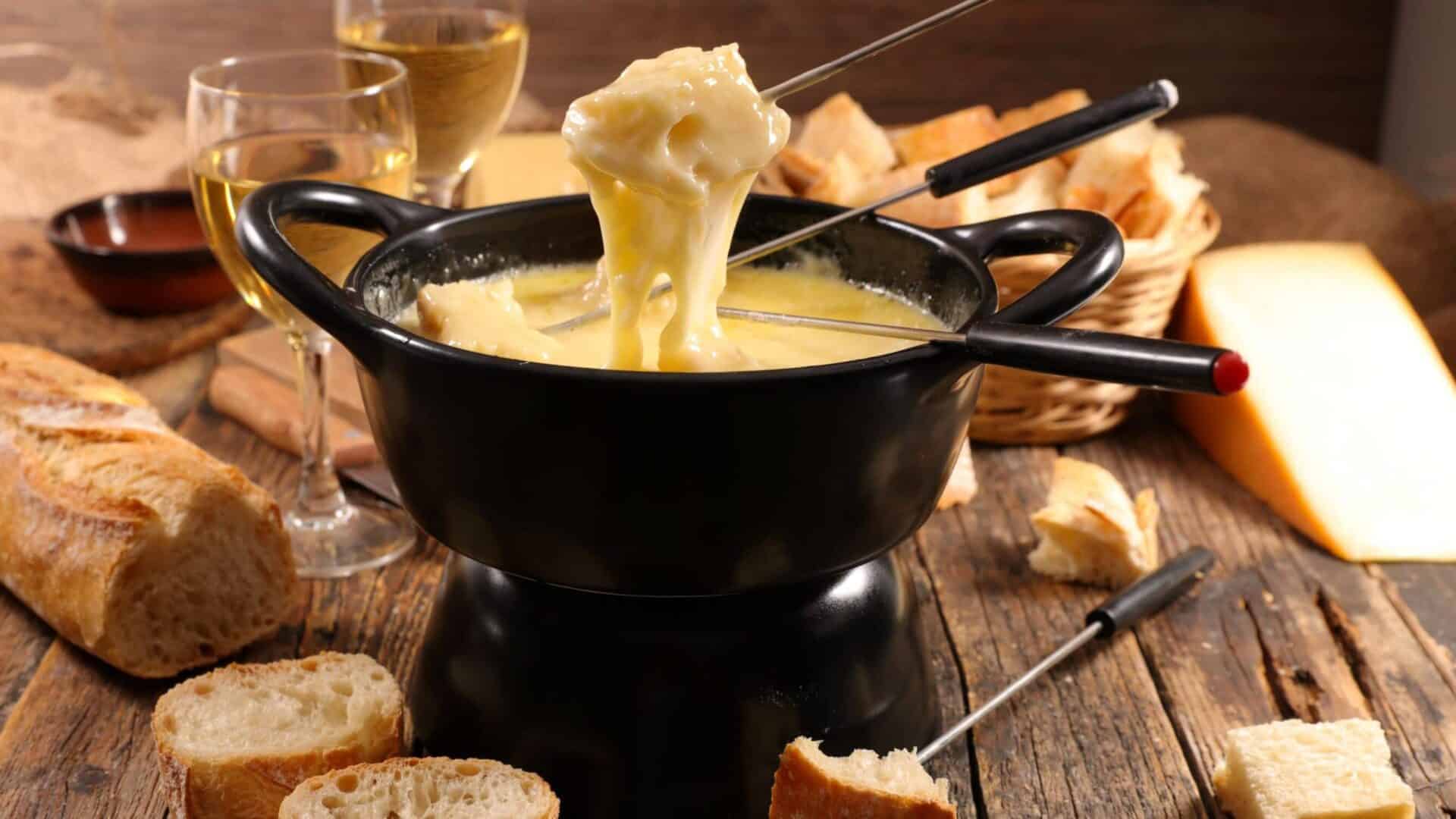 Black fondue pot with melted cheese and dipped bread with wine and more bread and cheese in the background