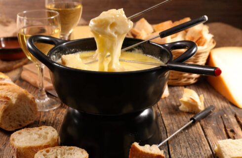 Black fondue pot with melted cheese and dipped bread with wine and more bread and cheese in the background
