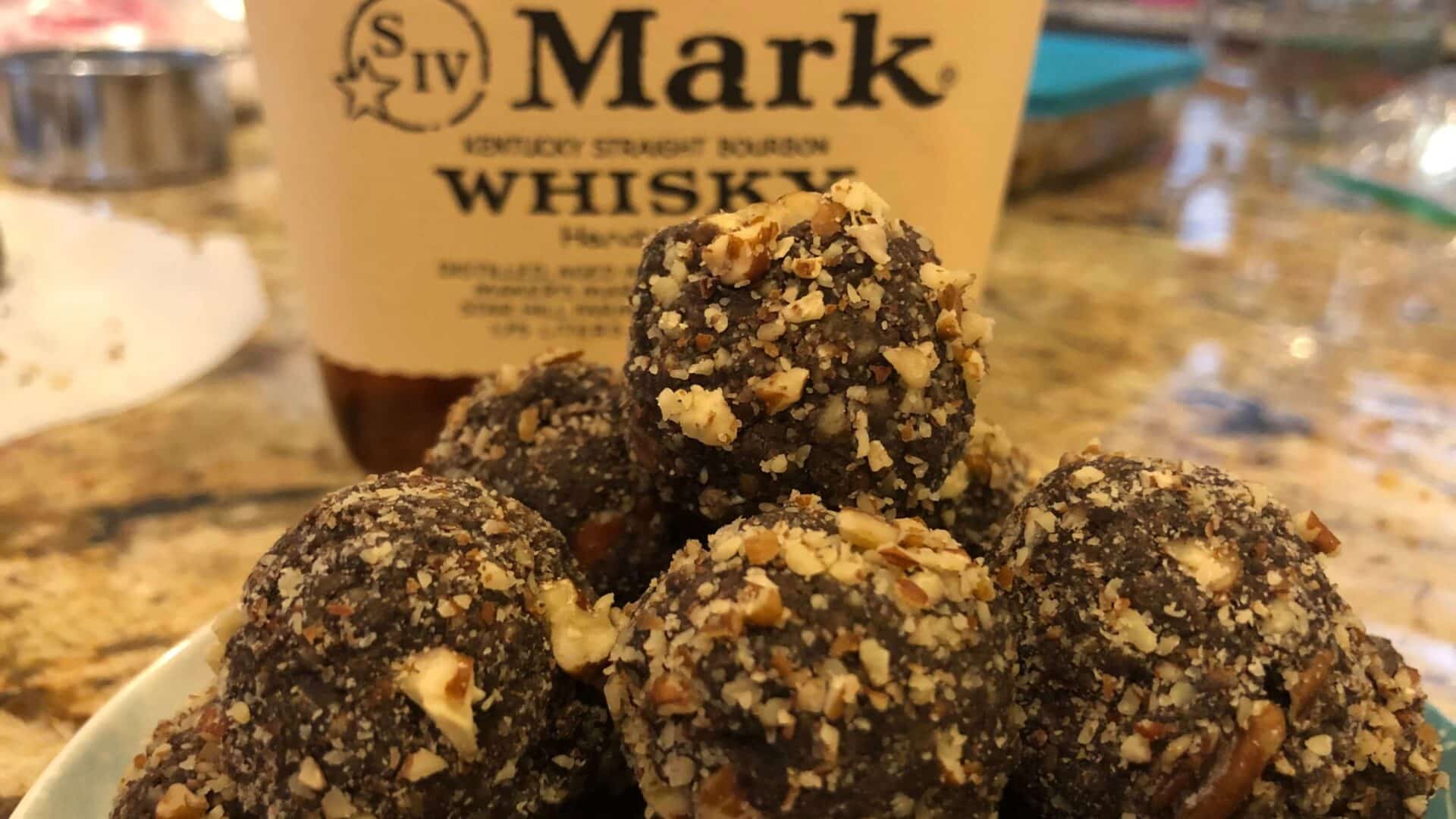 Round dark brown dough balls coated with chopped pecans and a bottle of bourbon whiskey in the background