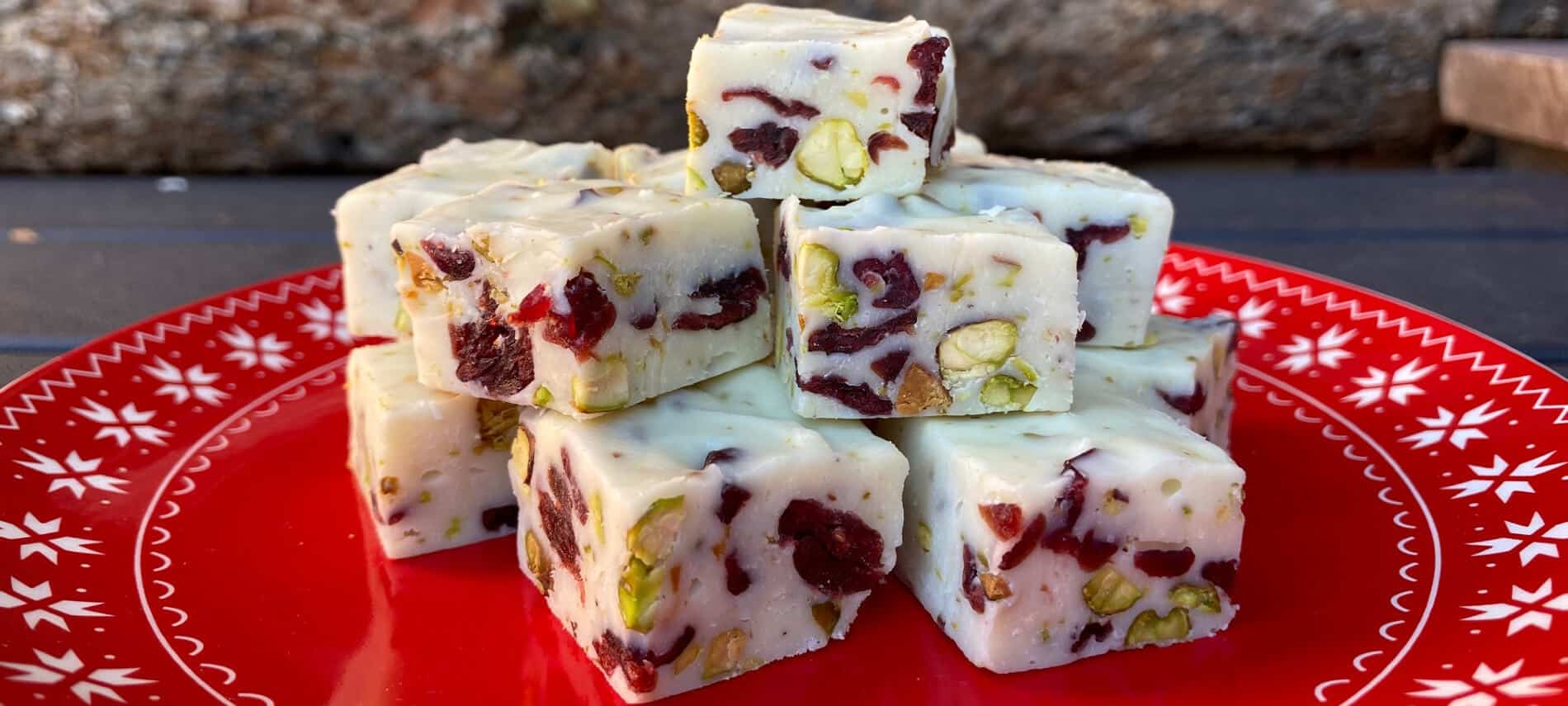squares of white chocolate fudge with red cranberries and green pistachios on a red plate with white snowflakes
