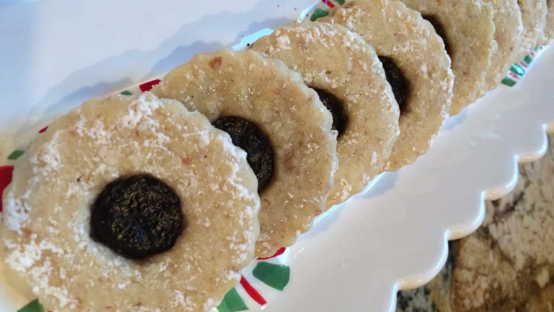 Round nutty cookies with cut out in the middle filled with a reddish purple jam, sprinkled with powdered sugar