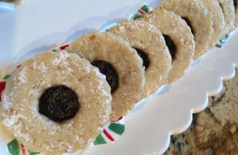 Round nutty cookies with cut out in the middle filled with a reddish purple jam, sprinkled with powdered sugar