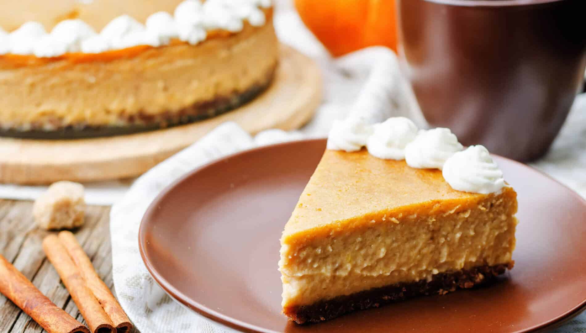 Orange pumpkin cheesecake with a brown gingerbread crust and dolloped with whipped cream on and brown plate and pumpkins as decorations