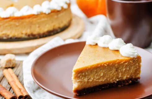 Orange pumpkin cheesecake with a brown gingerbread crust and dolloped with whipped cream on and brown plate and pumpkins as decorations