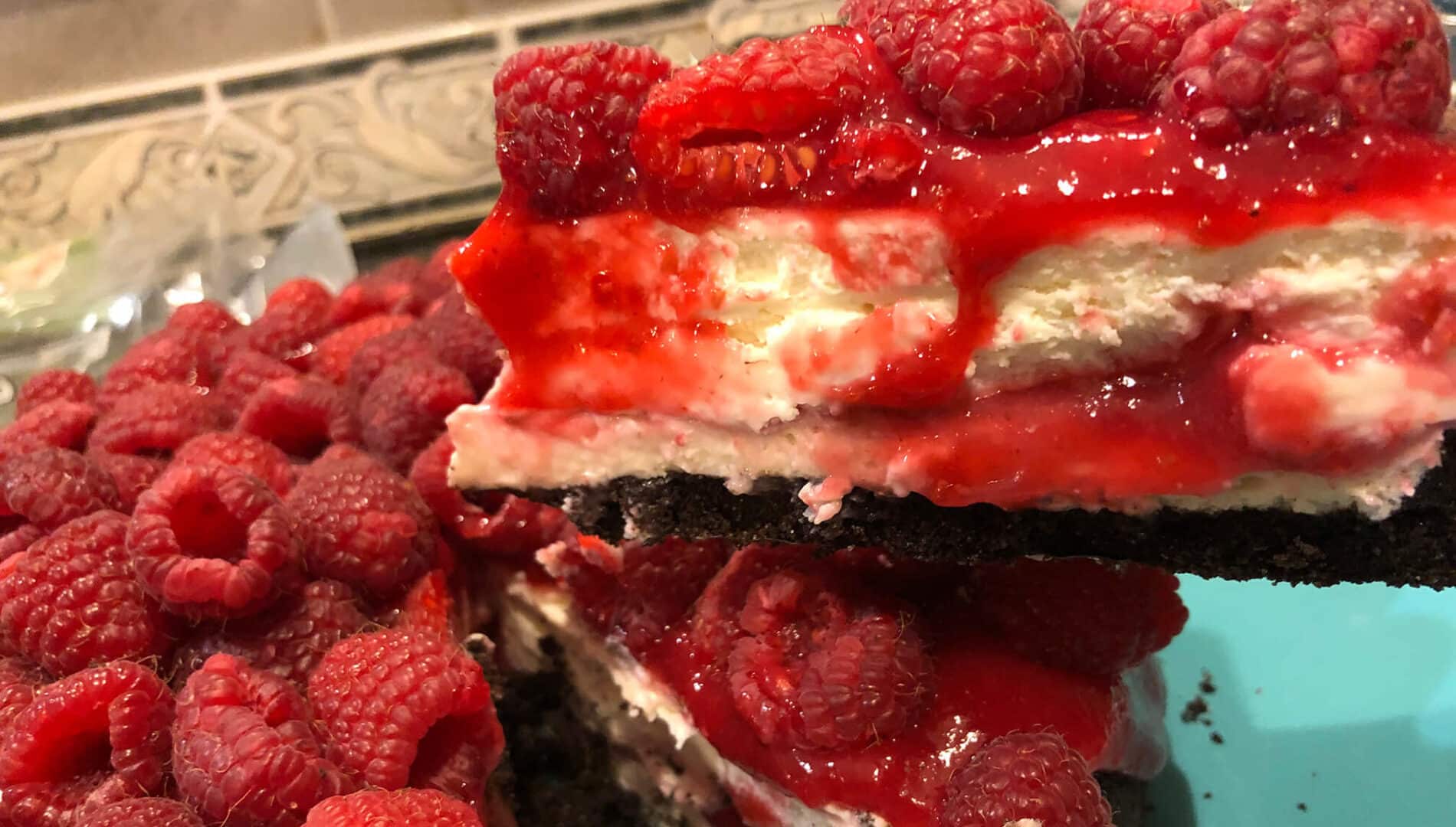 Creamy White Cheesecake with a red raspberry sauce topped with fresh strawberries, under a dark brown chocolate crust