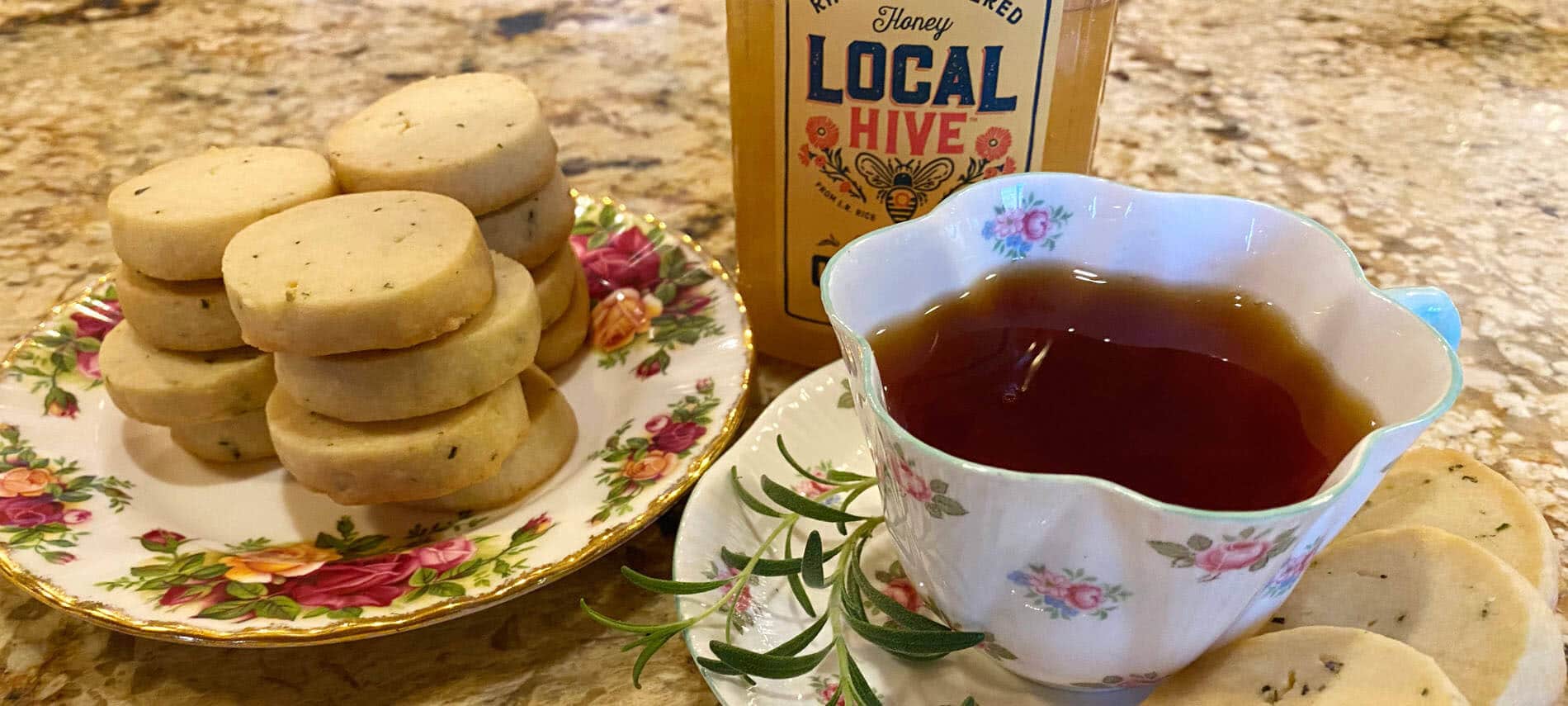 Round shortbread cookies with specs of rosemary along with a cup of tea a a jar of honey