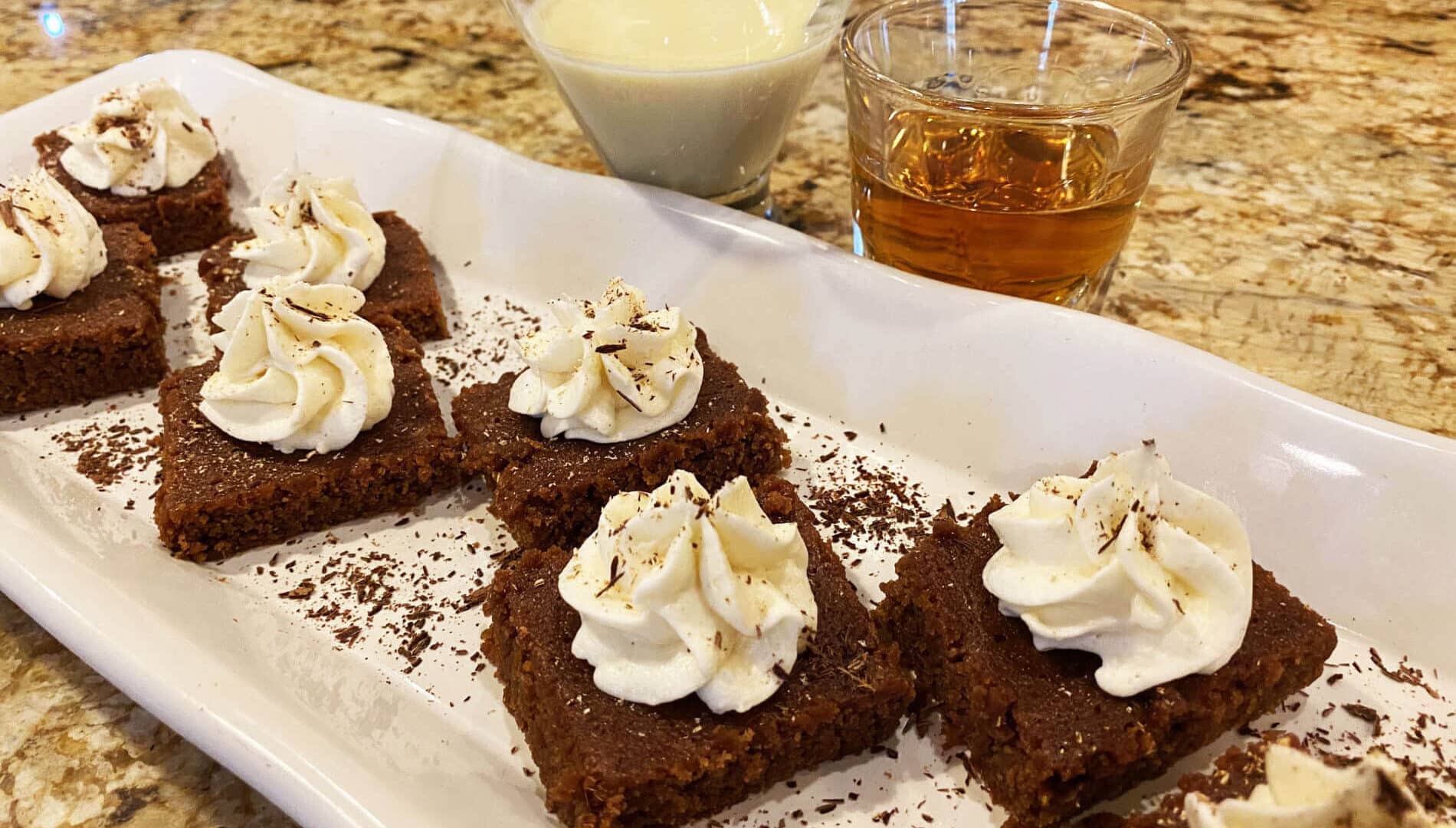 white plate with square brown brownies topped with white whipped cream and sprinkled with shaved chocolate, and glasses of white eggnog and golden bourbon