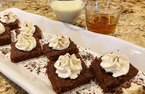 white plate with square brown brownies topped with white whipped cream and sprinkled with shaved chocolate, and glasses of white eggnog and golden bourbon