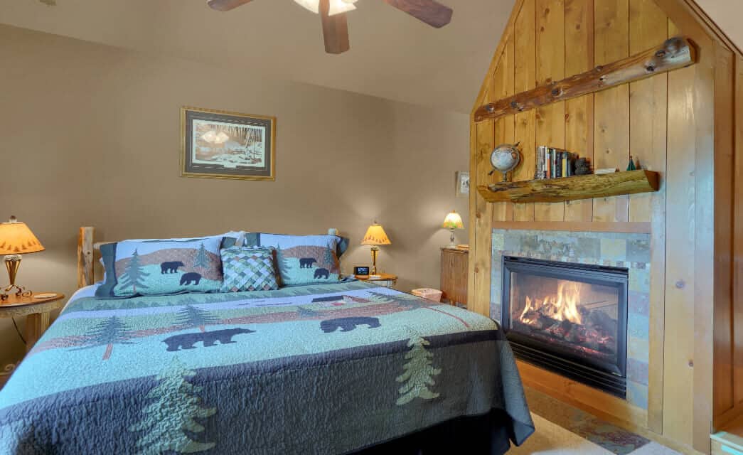 a king bed with a blue quilt with pine trees and bears, and a fireplace within a wood mantle next to the bed.
