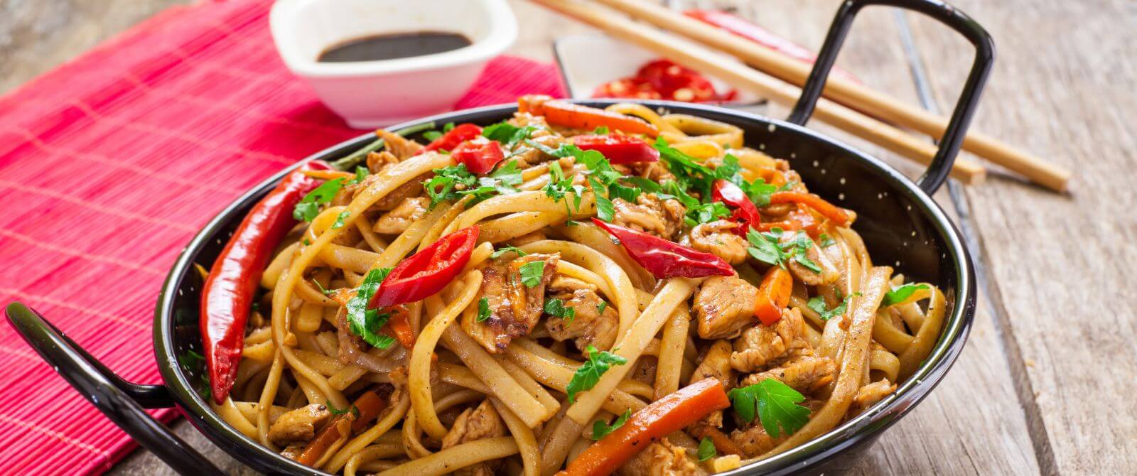 a wok full of noodles, peppers and vegetables with chopsticks and a bowl of soy sauce on the side