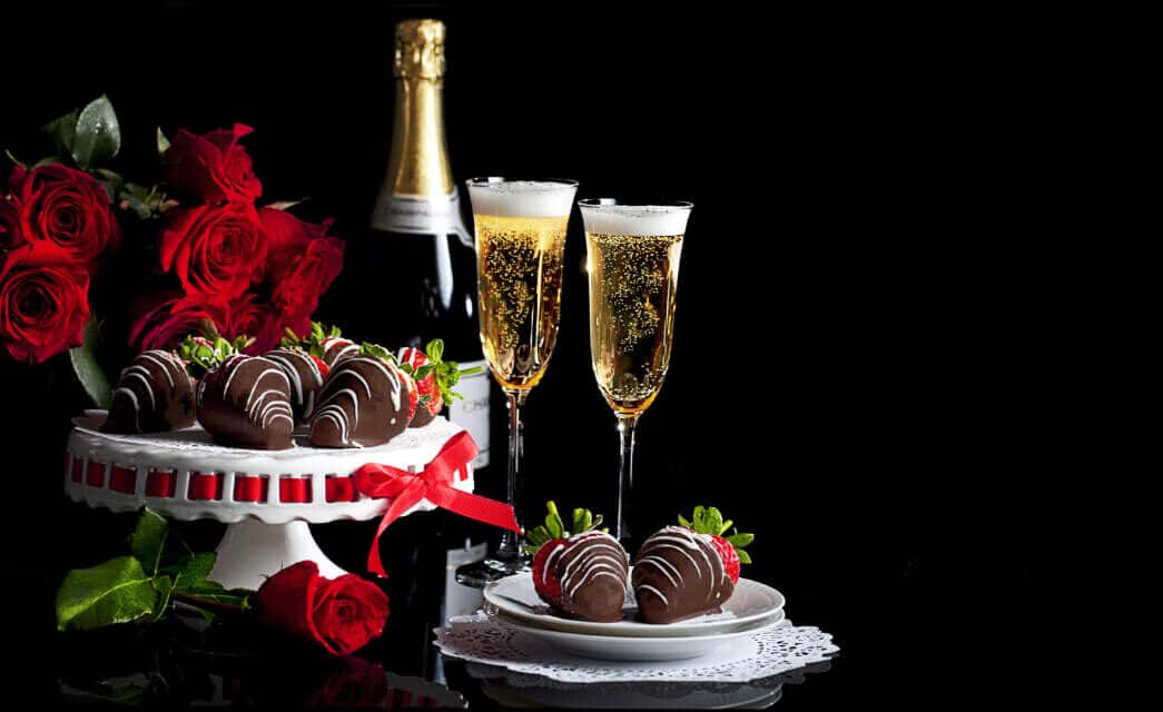 a plate of chocolate covered strawberries with two glasses of champane, a bottle of unopened champagne, and a bouquet of red roses