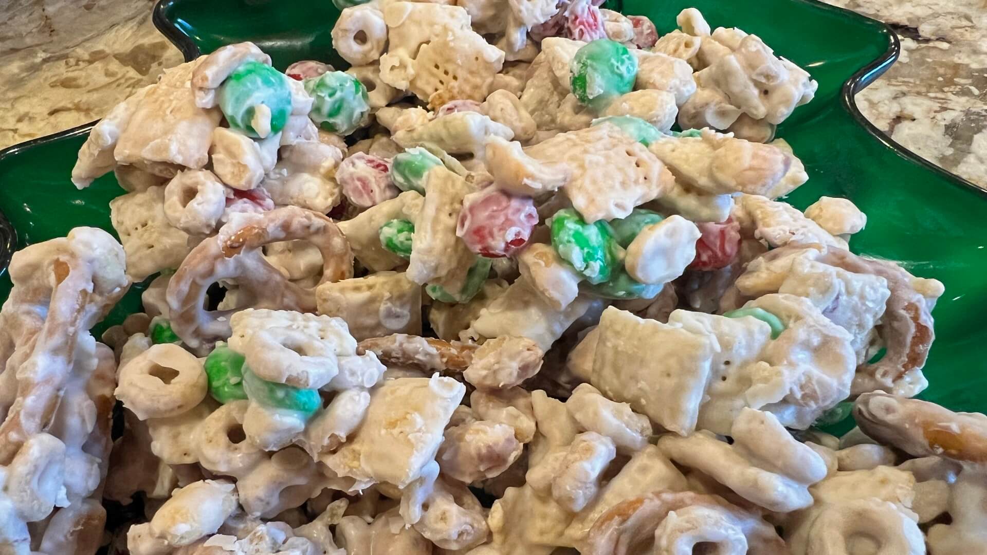 A Christmas candy of Chex® and Cheerios® cereals, peanuts, pretzels, green and red M&Ms®, coated with white chocolate on a green tray.