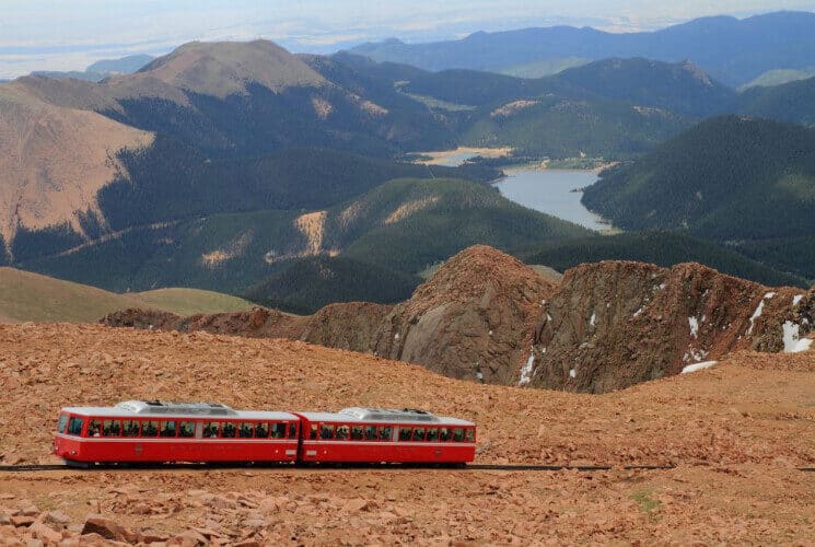 a red train going up a high mountain pass with mountains and a lake in the background