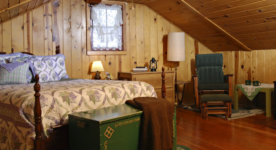 knotty pine wood panelled walls and pitched roofceiling with one window with lace window treatment, large 4 wood bed with cream, soft green and purple quilt, antique green chest with brown throw blanket in front and green and wood chair with ottaman
