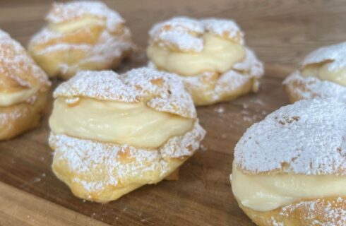 A wooden board with six large cream puffs filled with pastry cream and sprinkled with powdered sugar
