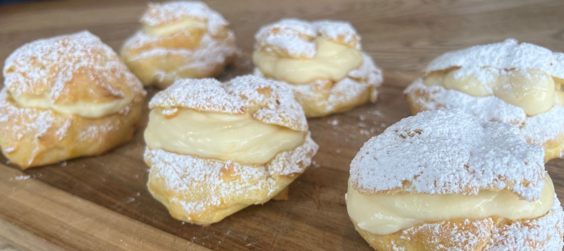 A wooden board with six large cream puffs filled with pastry cream and sprinkled with powdered sugar