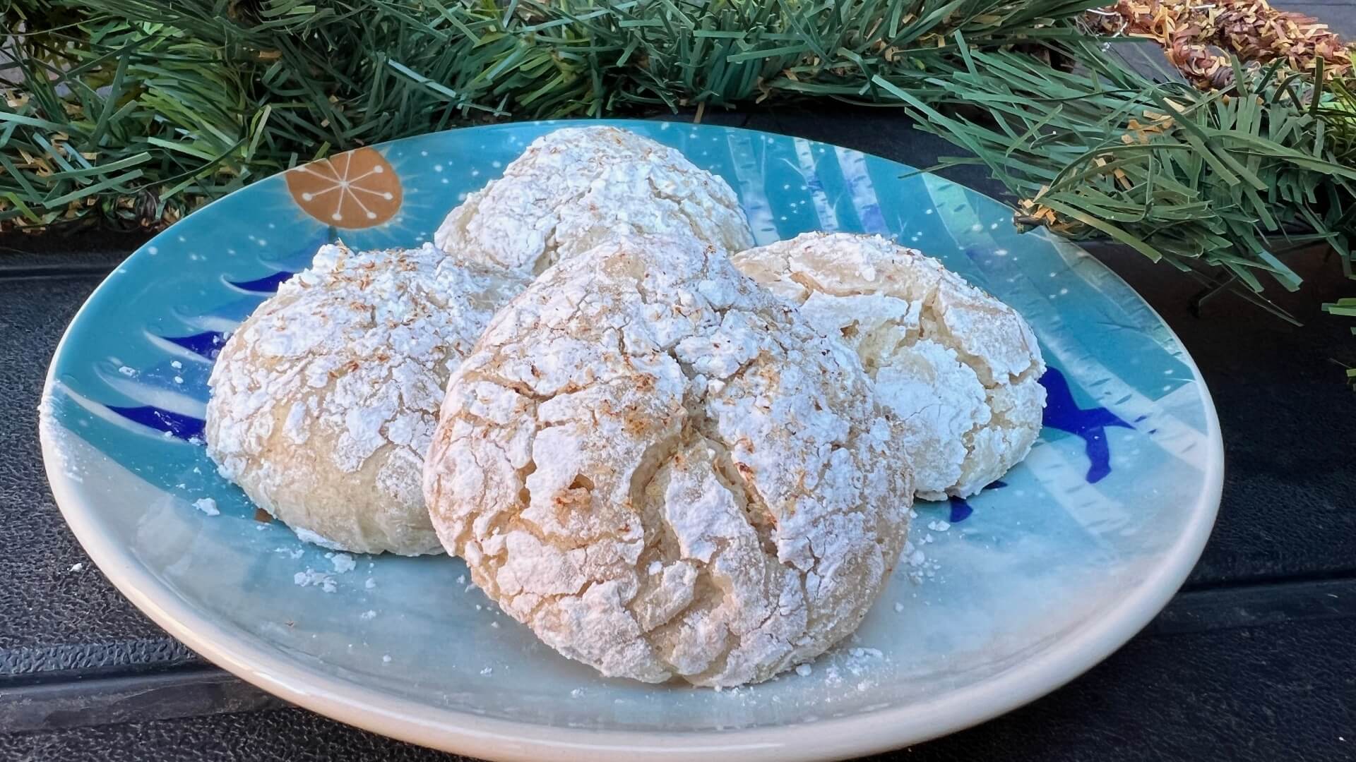 Round cookies baked in powdered sugar with crackles on the top, on a blue plate with pine garland in the background.