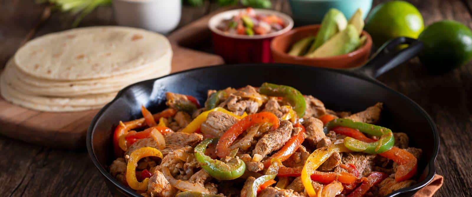 a sizzling plate of steak fajitas with red and green peppers, onions, and a plate of tortillas and bowls of salsa and sliced avocados in the background.