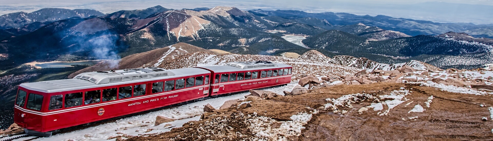 red train cars going up a mountain with snow capped mountains around