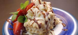 White and blue plate topped with delicious dessert drizzled with brown and white sauce and red strawberries