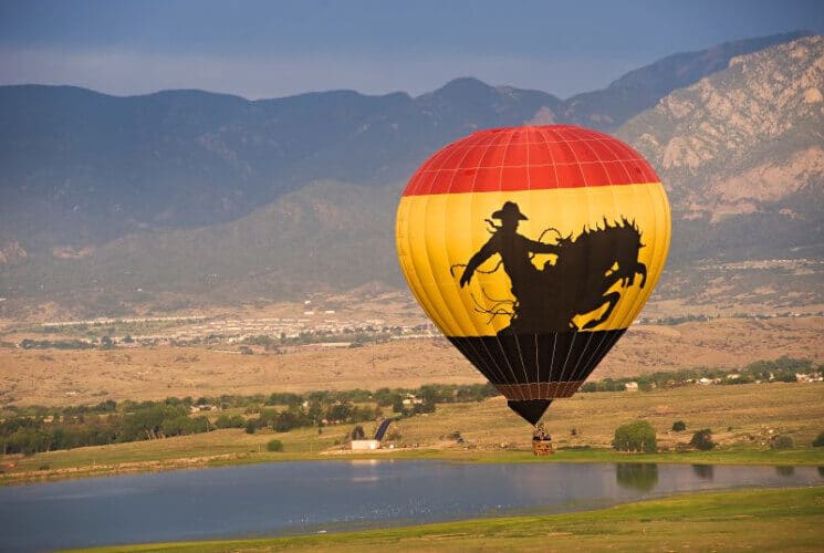 a hot air balloon flying high above the ground with green grass, lakes and mountains below