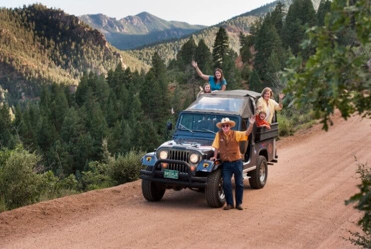 people standing by a jeep on a dirt road with pine tees and majestic mountains behind them