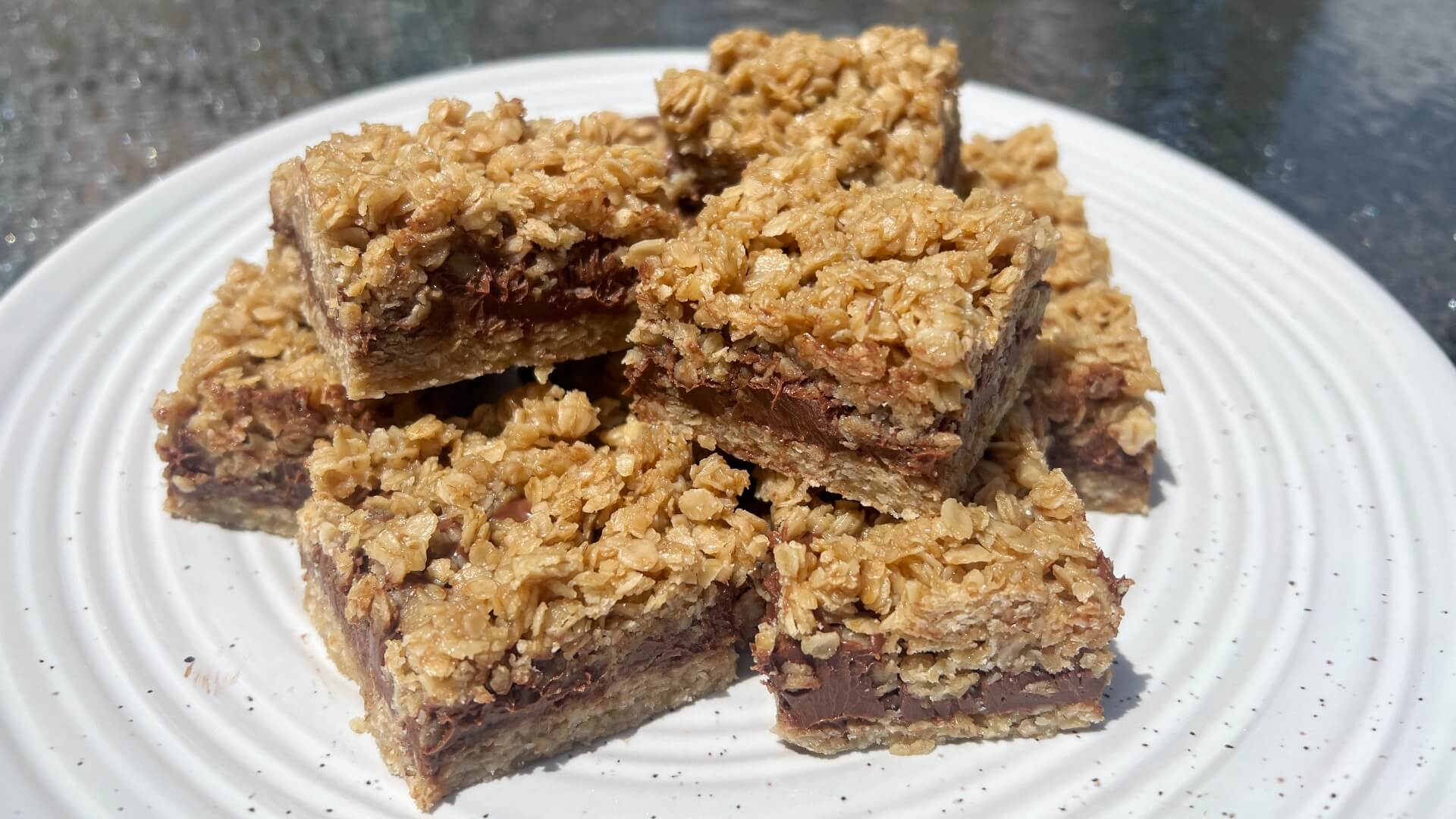 A white speckled plate with square oat cookie bars filled with a chocolate fudgy filling.