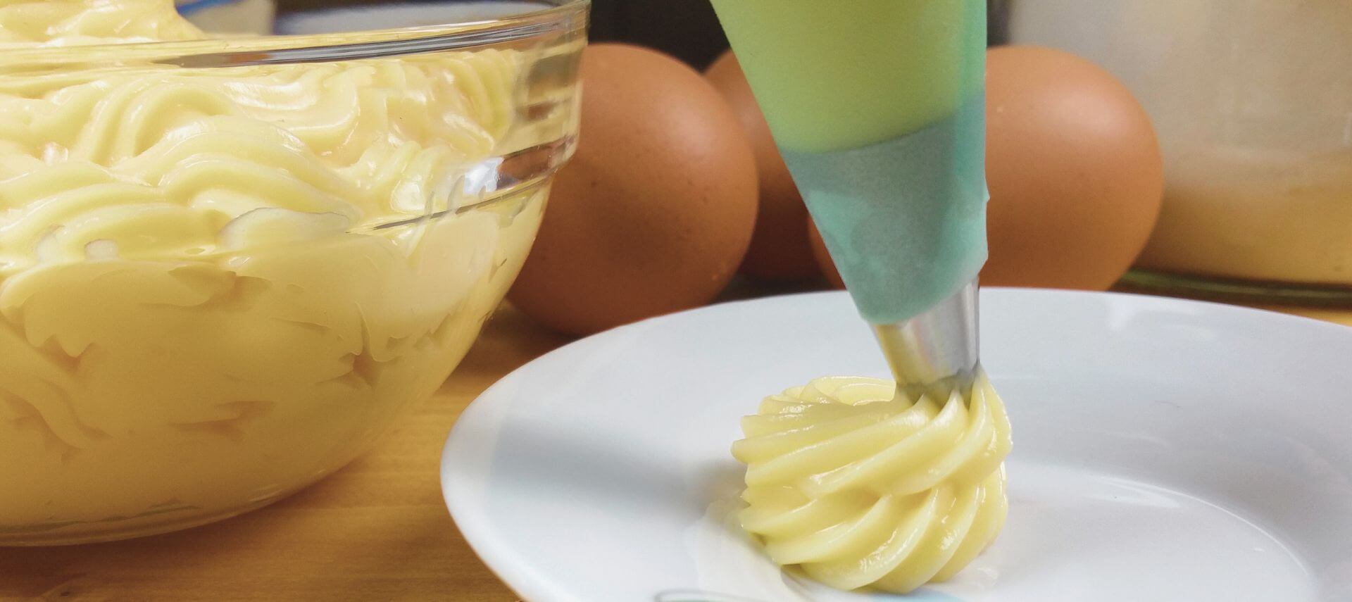 A bowl of pastry cream and some in a piping bag being piped onto a white plate, with 2 brown eggs in the background.