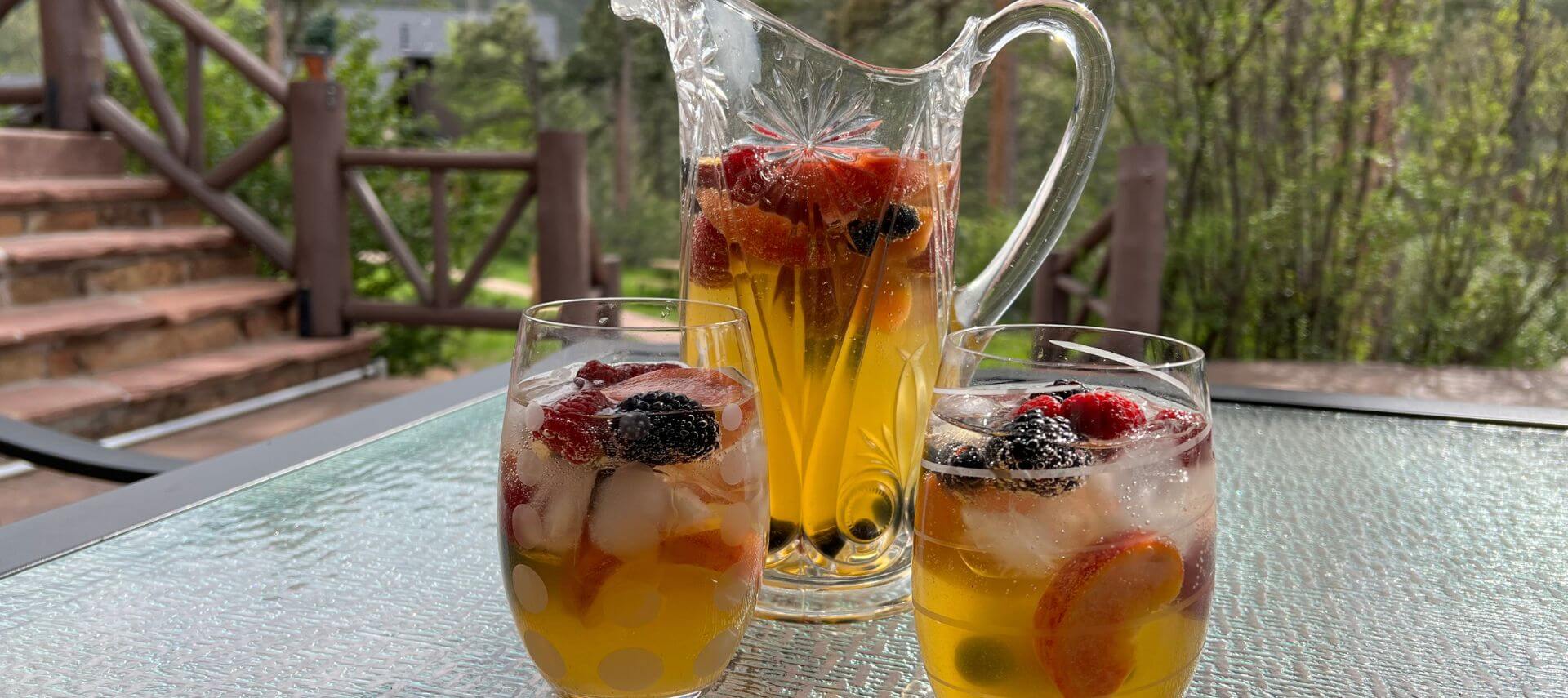 Two clear stemless wine glasses with a white wine with peaches and berries, with a pitcher of the sangria behind the glasses on a table outside with steps and green trees and mountains in the background.