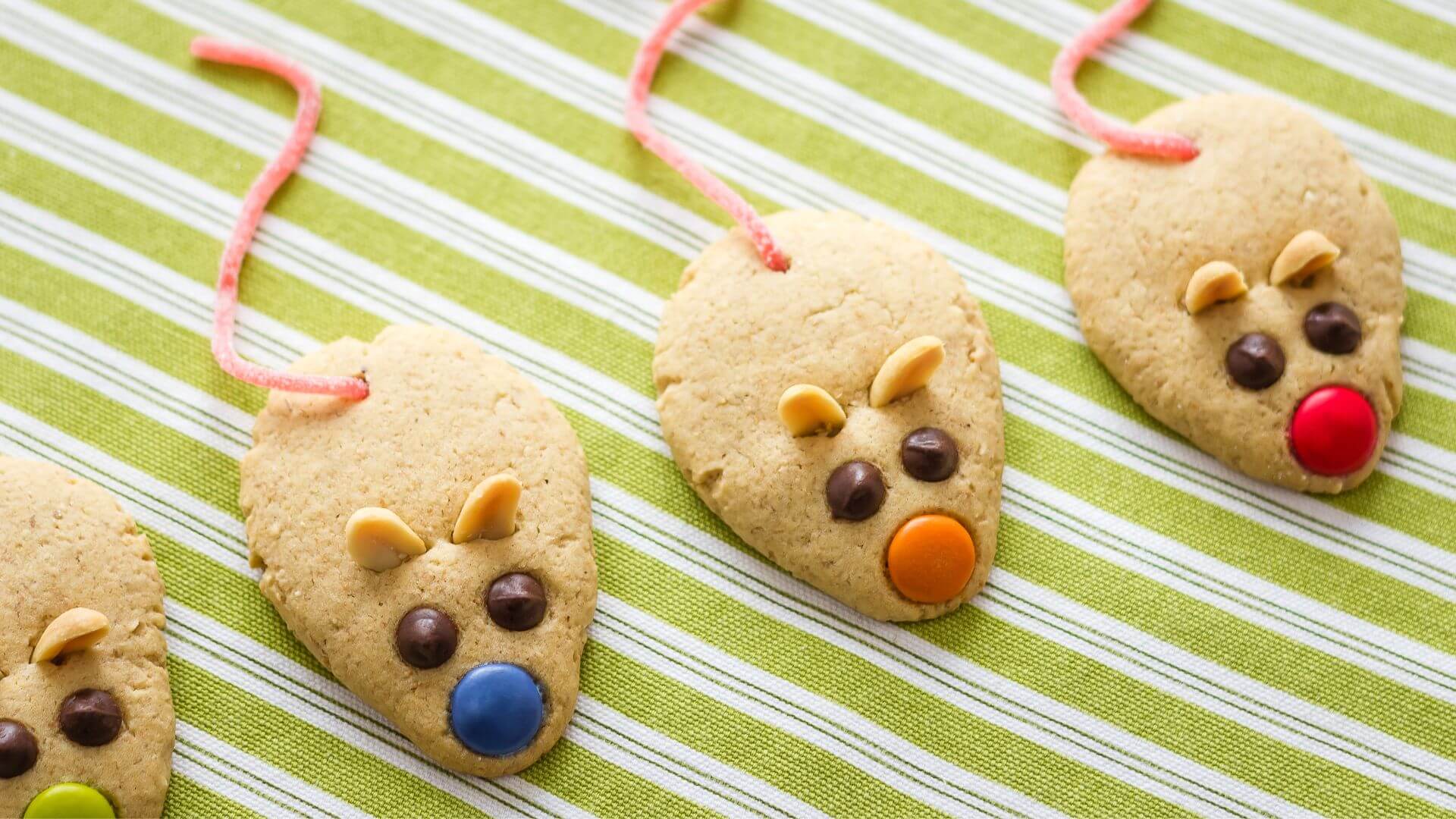 peanut butter cookies shaped like mice with mini chocolate chips and m&m's, peanut ears, and candy tails on a green and white striped cloth