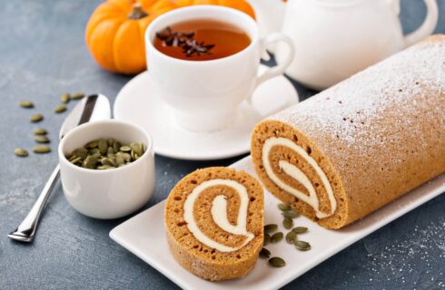 A delicate pumpkin roll with a creamy filling next to a cup of tea, pumpkin seeds and a teapot