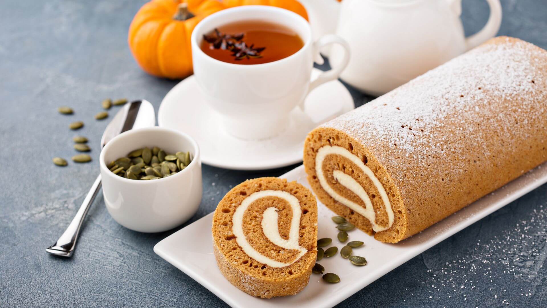 A delicate pumpkin roll with a creamy filling next to a cup of tea, pumpkin seeds and a teapot