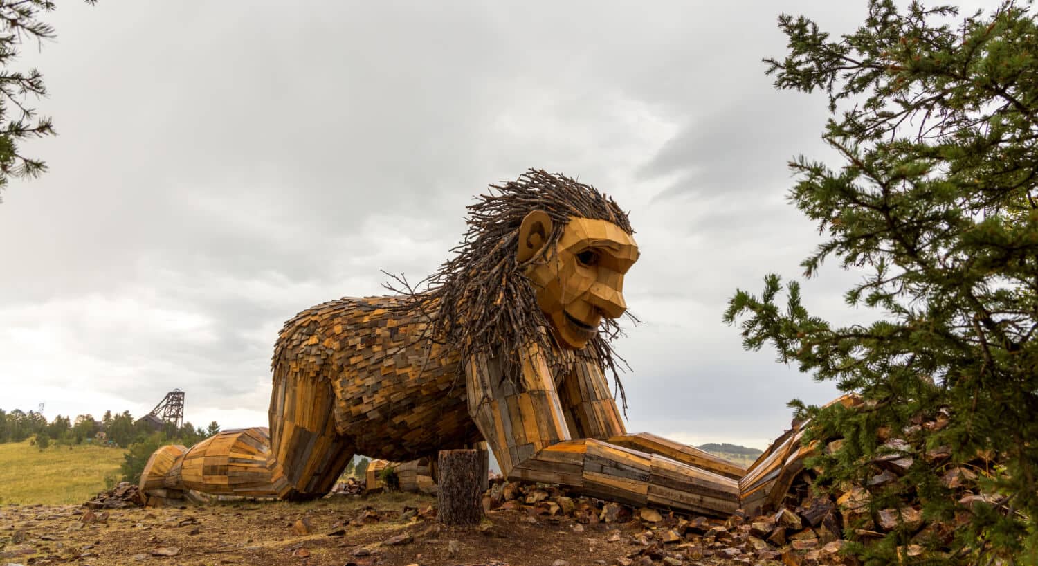 A large wood troll named Rita the Rock Planter made out of recycled wood kneeling and placing rocks in front of pine trees and a gold mining tower in the background.