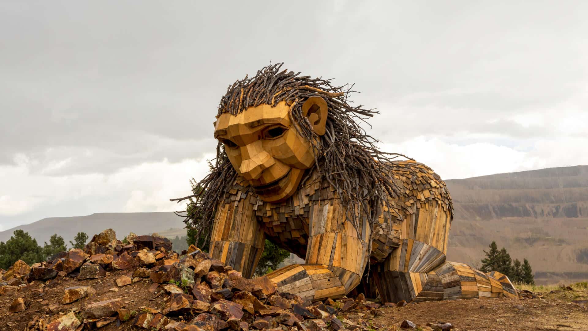 A large wood troll named Rita the Rock Planter made out of recycled wood kneeling in front of mountains.