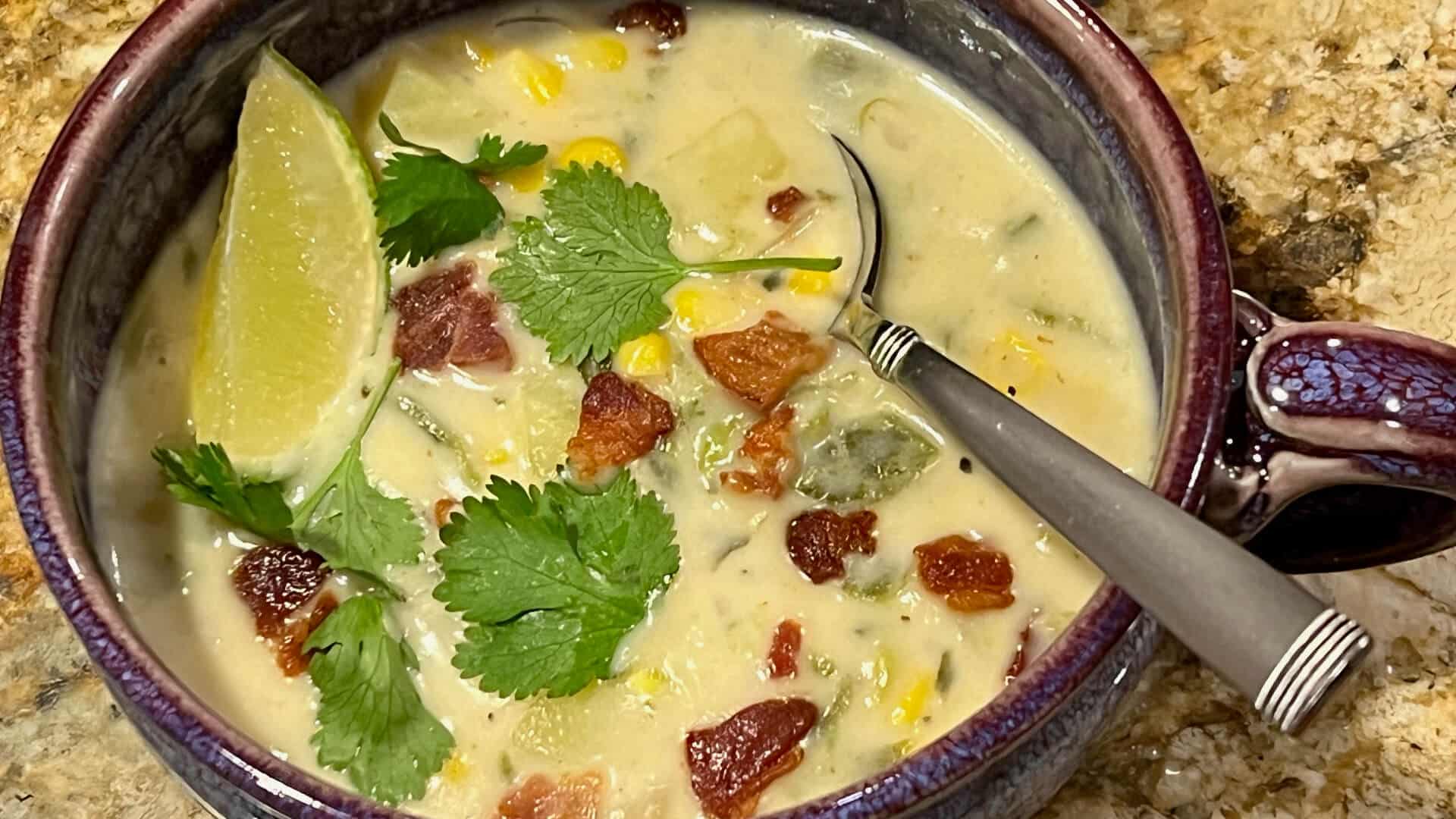 a purple bowl of creamy cheese soup with pieces of roasted poblano peppers, potatoes, and corn, garnished with crispy bacon pieces, cilantro and a lime wedge
