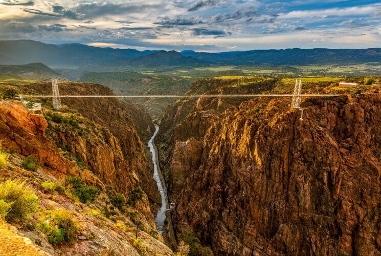 a suspension bridge high above a rive in between two walls of red rocks and sweeping mountains in the distance