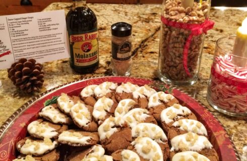 A tray with gingersnap cookies dipped in white chocolate and dotted with pieces of crystallized ginger, with a recipe, molasses, ginger, nuts and a candle in the background