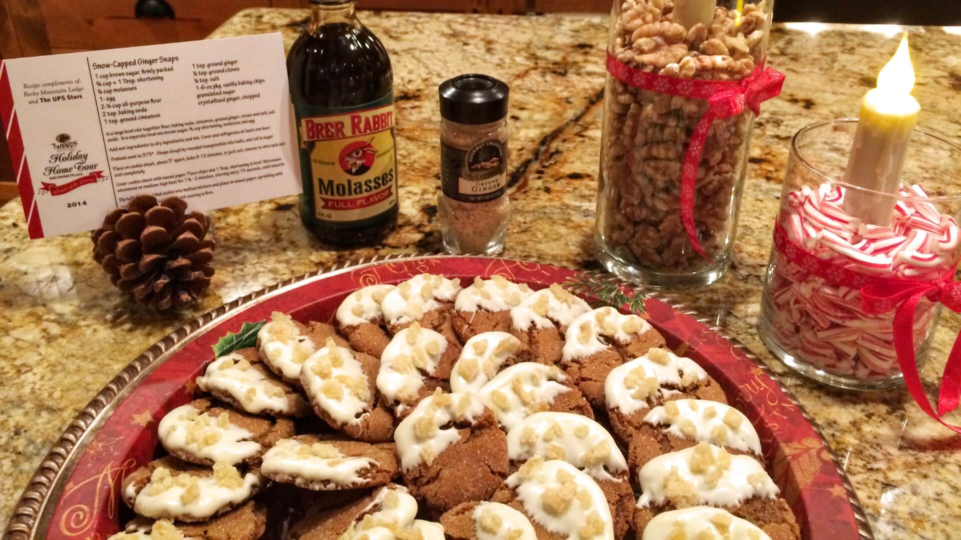 A tray with gingersnap cookies dipped in white chocolate and dotted with pieces of crystallized ginger, with a recipe, molasses, ginger, nuts and a candle in the background
