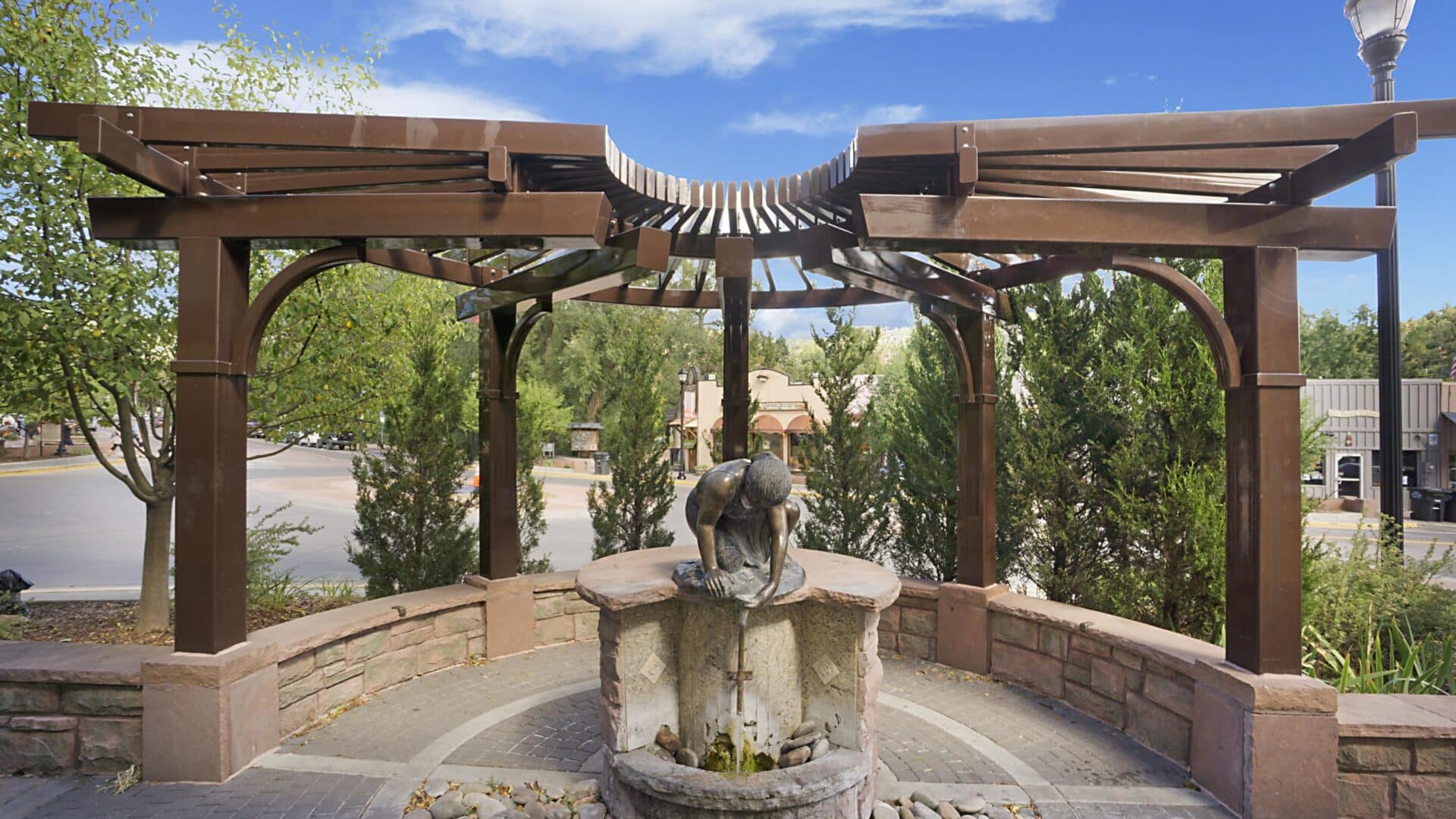 Mineral spring with bronze statue above it catching water in a half circle of brick and wood with blue skies and clouds, and trees and bushes around