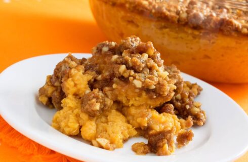 a white plate with mashed orange sweet potatoes with a streusel topping with pecans on top and a bowl of the casserole in the background