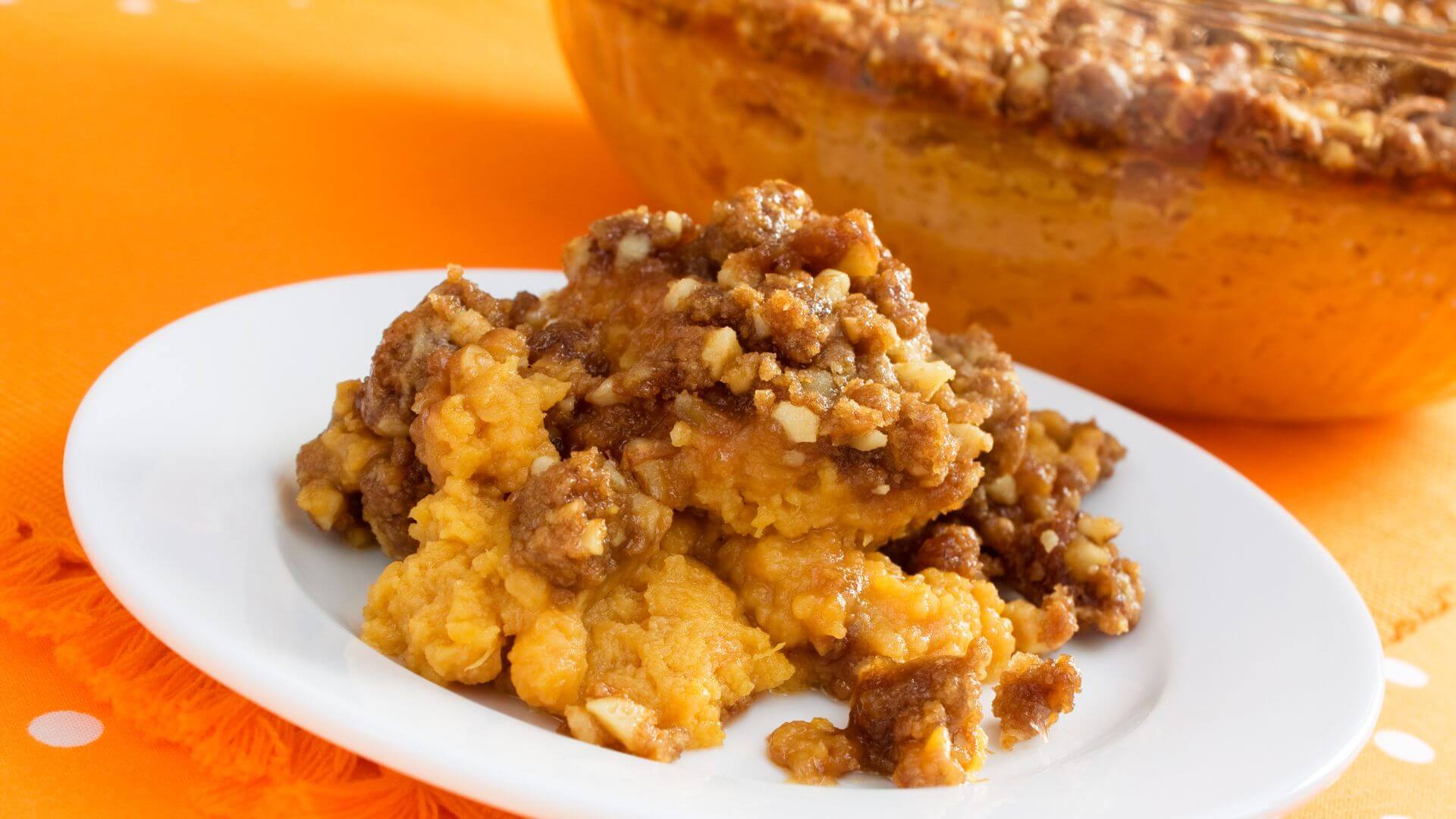 a white plate with mashed orange sweet potatoes with a streusel topping with pecans on top and a bowl of the casserole in the background