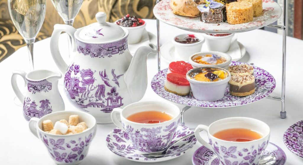 A table set for tea with a purple and white tea set, and a tiered platter with finger foods.